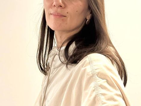Woman with long brown hair wearing a light blouse standing against a light background.