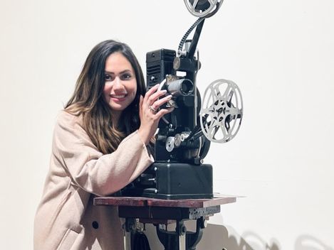 Person mit langem Haar und hellem Mantel bedient einen alten Filmprojektor mit zwei Filmrollen auf einem Tisch.