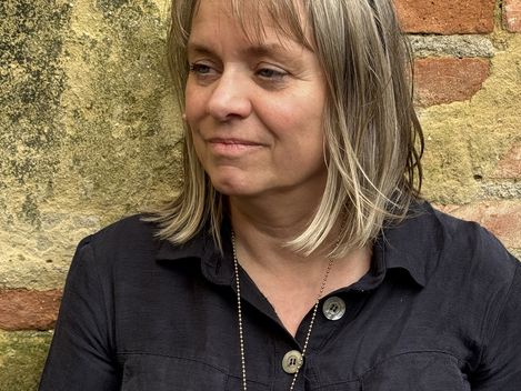 Frau mit schulterlangem Haar, schwarzem Hemd mit Knöpfen und Brille auf dem Kopf vor einer gemauerten Wand.