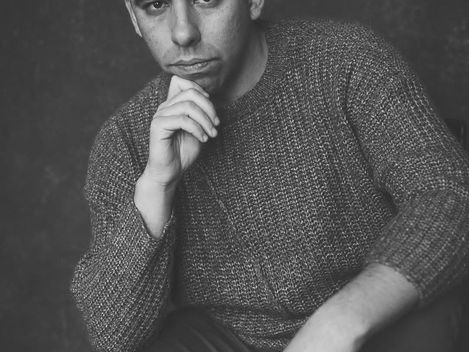 Person wearing a beanie and sweater sitting with one hand on the chin and the other on the knee.