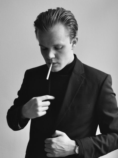 Black-and-white portrait of a man in a suit jacket holding the jacket with one hand and pointing to his chest with the other.