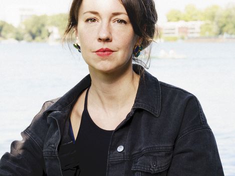Person with hair tied up wearing a black denim jacket and black top standing in front of a body of water with trees and buildings in the background.