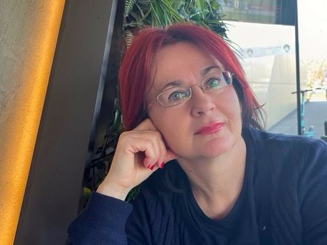 Woman with red hair and glasses sitting at a table, resting her head on her hand and looking forward.