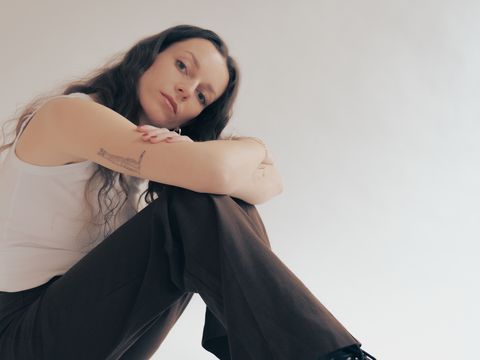 Person with long hair sitting with bent legs and crossed arms against a light background