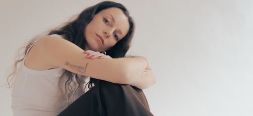 Person with long hair sitting with bent legs and crossed arms against a light background