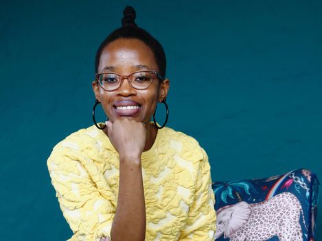 Person with hair in a top bun wearing a yellow textured top sitting on a chair with blue leopard print and an elephant-patterned cushion against a blue background.
