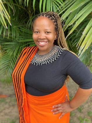 Woman with braided hair wearing a black top, an orange wrap skirt, and an orange-and-black scarf standing in front of green palm leaves with one hand on her hip.