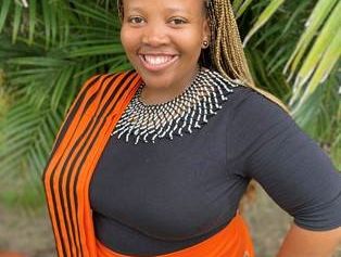 Woman with braided hair wearing a black top, an orange wrap skirt, and an orange-and-black scarf standing in front of green palm leaves with one hand on her hip.