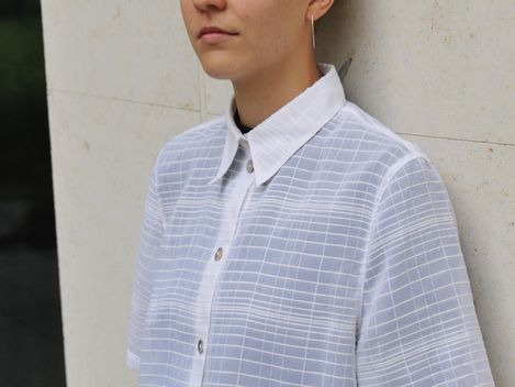 Person with short blond hair leaning against a light-colored wall wearing a short-sleeved checkered shirt with a white collar.