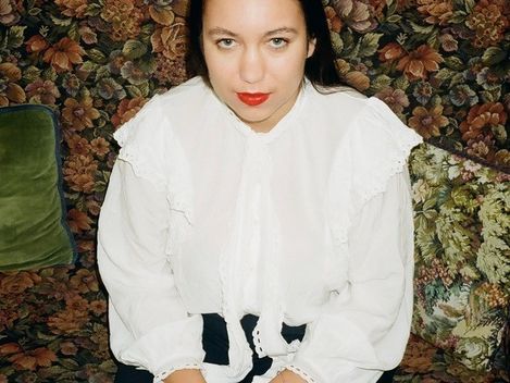 Person sitting on a floral-patterned sofa wearing a white blouse with ruffles and dark pants with hands folded in lap.