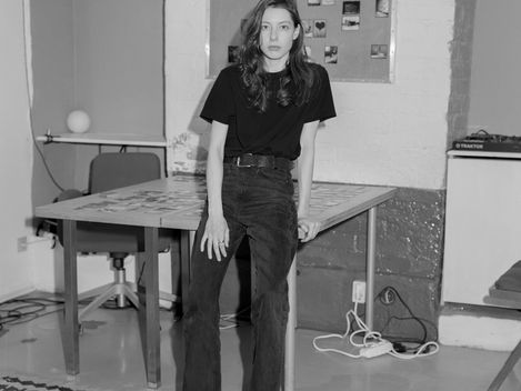 Person with shoulder-length hair wearing a black t-shirt and pants leaning against a table in a room with a rug and bulletin board in the background.