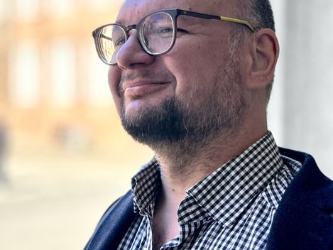 Porträt eines Mannes mit Brille und Bart, aufgenommen im Freien vor unscharfem architektonischem Hintergrund.