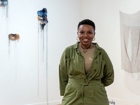 Person with short hair and hoop earrings wearing an olive green jacket standing in front of a white wall with abstract artworks.