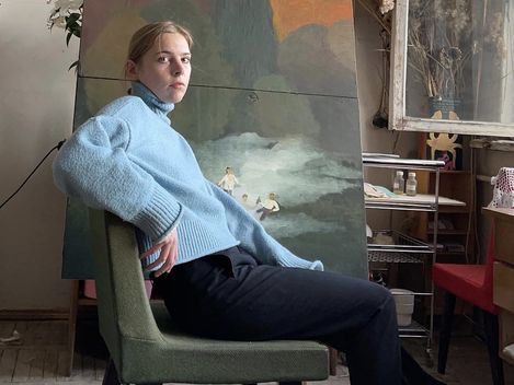 Person wearing a light blue sweater and dark pants sitting sideways on a chair in front of a large painting with abstract shapes and figures.