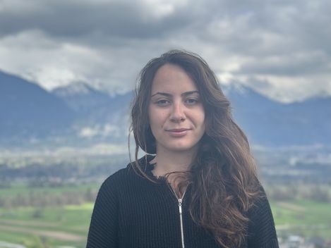 Person with long hair wearing a black sweater standing in front of a landscape with mountains and cloudy sky.