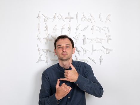 Man in dark shirt in front of white wall with abstract white shapes making hand signs.