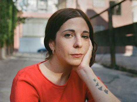 Portrait of a woman in a red top resting her face on her hand while looking directly at the camera.