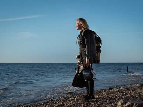 Person standing on a rocky shore wearing scuba diving gear under a clear sky.