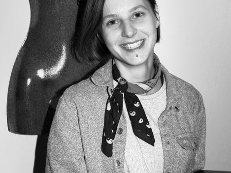 Person with shoulder-length hair wearing a scarf and shirt sitting in front of a black female mannequin torso.