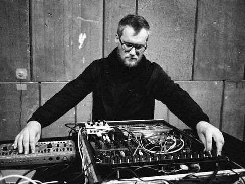 Person sitting at a table with electronic music equipment and mixer in front of a concrete wall