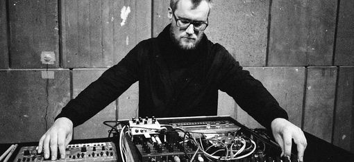 Person sitting at a table with electronic music equipment and mixer in front of a concrete wall