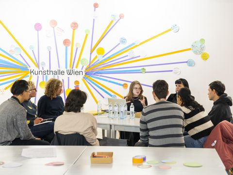 A group of people are sitting around a table listening to a person who is explaining something.