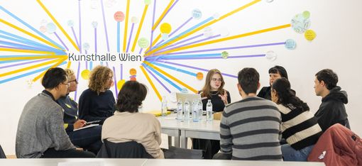 A group of people are sitting around a table listening to a person who is explaining something.