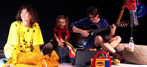 The image features a man and two children sitting on the floor, playing with toys. The man is holding a guitar, playing music for the children.