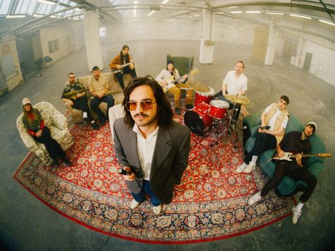 Eine neunköpfige Band sitzt und steht mit Instrumenten auf einem großen gemusterten Teppich in einer Industriehalle. Im Vordergrund steht ein Mann mit Mikrofon und Sonnenbrille.