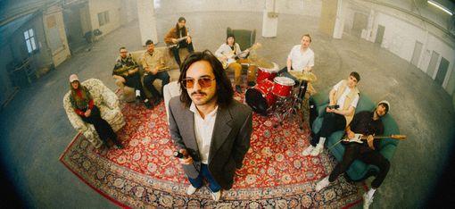 Eine neunköpfige Band sitzt und steht mit Instrumenten auf einem großen gemusterten Teppich in einer Industriehalle. Im Vordergrund steht ein Mann mit Mikrofon und Sonnenbrille.