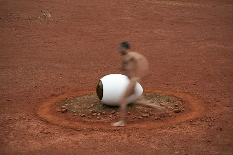 Blurred image of a naked man running past a large white eye-shaped object on red ground