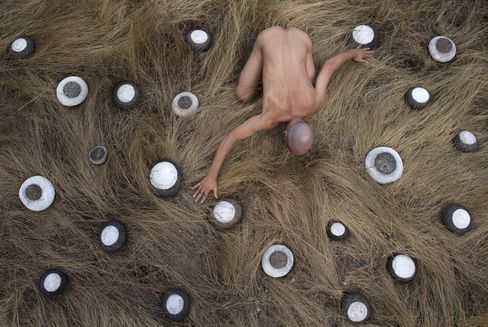 Top view of a naked man lying with outstretched arms on a floor covered with long straw-like material surrounded by multiple round objects with white centers and dark edges