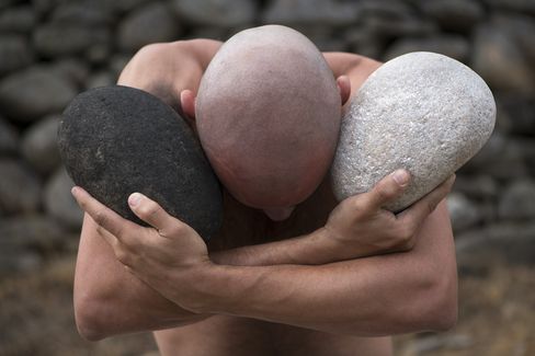 Upper body of a shirtless man with bowed head holding two large stones on his shoulders