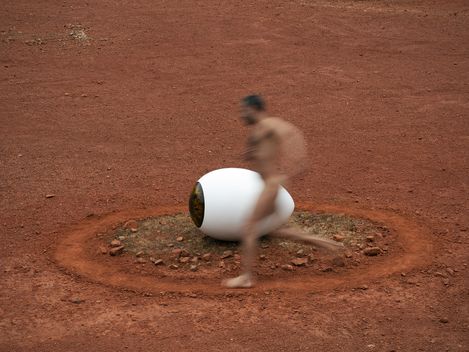Blurred image of a naked man running past a large white eye-shaped object on red ground