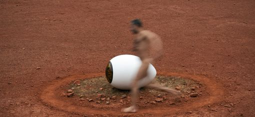 Blurred image of a naked man running past a large white eye-shaped object on red ground