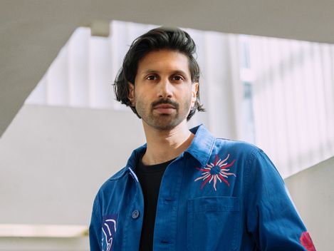Person with medium-length dark hair wearing a blue shirt with colorful embroidery standing in a bright modern interior with stairs and railing.