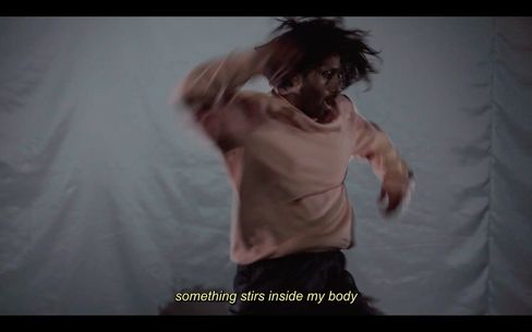 Person with long hair and sweater in motion against a plain background with subtitle "something stirs inside my body".