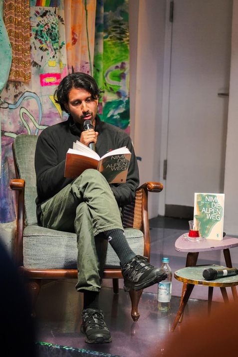 Person sitting in an armchair holding a book titled "Nimm den Alpenweg" and speaking into a microphone with colorful wall art in the background.