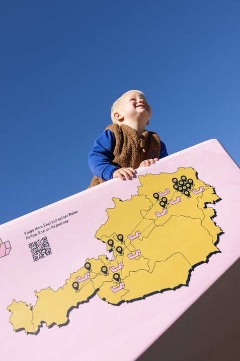 Ein lachendes Kind auf einem rosa Enzi mit Österreichkarte und Stationen der Enzi-Tour vor blauem Himmel.