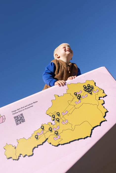 Ein lachendes Kind auf einem rosa Enzi mit Österreichkarte und Stationen der Enzi-Tour vor blauem Himmel.