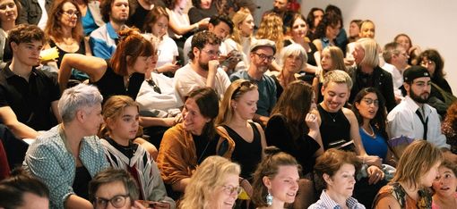 Große Zuschauermenge sitzt dicht gedrängt auf Stufen, einige lachen, reden und schauen nach vorne.