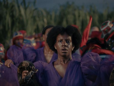 Frau mit Afro-Frisur in violettem Kleid hält eine Schildkröte, umgeben von Menschen in farbiger Kleidung und Kopftüchern vor einer grünen Pflanzenkulisse
