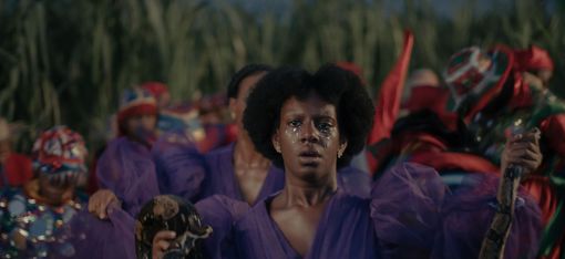 Frau mit Afro-Frisur in violettem Kleid hält eine Schildkröte, umgeben von Menschen in farbiger Kleidung und Kopftüchern vor einer grünen Pflanzenkulisse