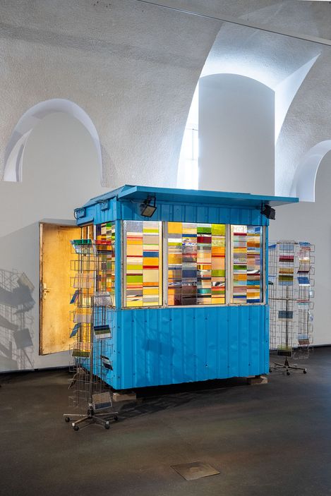 Blauer Kiosk mit farbigen Schubladen und zwei drehbaren Drahtgestellen in einem Raum mit hohen, gewölbten Fenstern