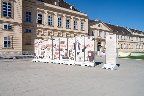 Mehrere bemalte Betonabsperrungen mit der Aufschrift 'WHAT ARE YOU WAITING FOR TODAY' vor einem historischen Gebäude mit gelber Fassade und Satteldach.