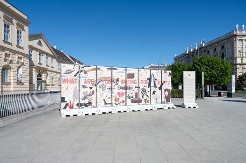 Große weiße Absperrwand mit bunten Graffiti und der Aufschrift 'WHAT ARE YOU WAITING FOR TODAY' auf einem großen Platz vor historischen Gebäuden bei klarem Himmel.