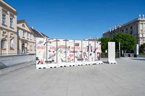 Große weiße Absperrwand mit bunten Graffiti und der Aufschrift 'WHAT ARE YOU WAITING FOR TODAY' auf einem großen Platz vor historischen Gebäuden bei klarem Himmel.