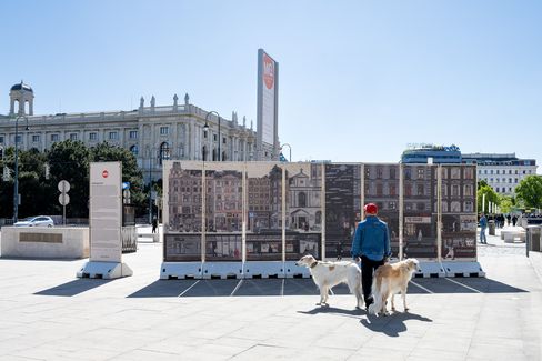 Person mit roter Mütze und blauer Jacke steht mit zwei Hunden vor einer Reihe von Informationstafeln auf einem Platz mit historischen Gebäuden im Hintergrund
