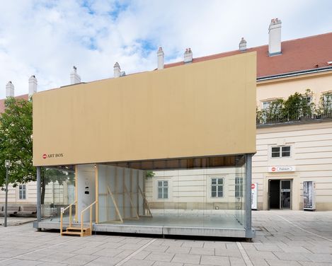Glascontainer mit beige verkleideter Oberseite und Holztreppe vor einem historischen Gebäude auf einem gepflasterten Platz