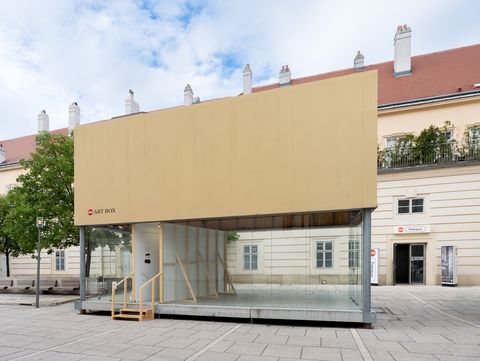 Glass container with beige-clad upper section and wooden stairs in front of a historic building on a paved square